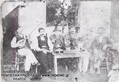 Ο Νίκος Μιλάνος (γνωστός ως Κολατσής) παίζει μπουζούκι στην παρέα του, στον Πορταριά του Πηλίου, το 1895. Στο παράθυρο ο γιός του Στέφανος, ντυμένος ναυτάκι. Το εικονιζόμενο μπουζούκι σώζεται σε άριστη κατάσταση